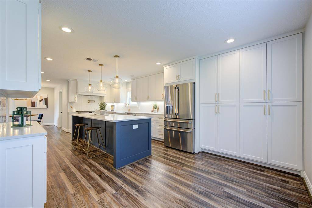 This spacious kitchen features modern design with sleek white cabinetry, a large island with seating, stainless steel appliances, and elegant pendant lighting. The wood flooring adds warmth, creating a welcoming and functional space for cooking and entertaining.