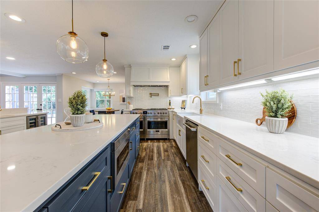This kitchen features modern design with white cabinetry, brass hardware, and a large island with navy blue base. It includes stainless steel appliances, pendant lighting, and wood-style flooring, creating a bright and spacious cooking area.