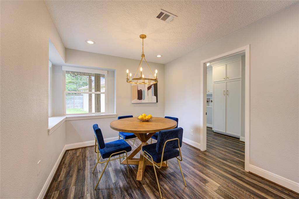 This cozy dining area features a round wooden table with blue velvet chairs, highlighted by a modern chandelier. It opens to a kitchen with ample cabinetry, and a large window offers natural light and views of the backyard.