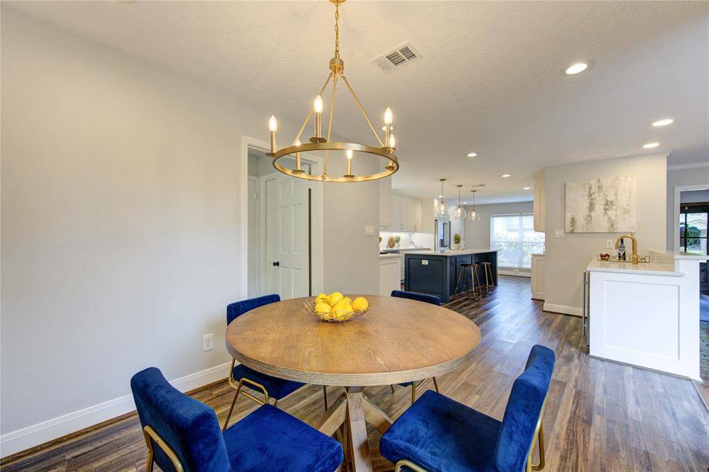 This inviting open-concept space features a modern kitchen with white cabinetry, a spacious island, and sleek countertops. The adjoining dining area includes a round table with elegant blue chairs and a stylish chandelier, all set on warm wood flooring, creating a welcoming atmosphere for gatherings.