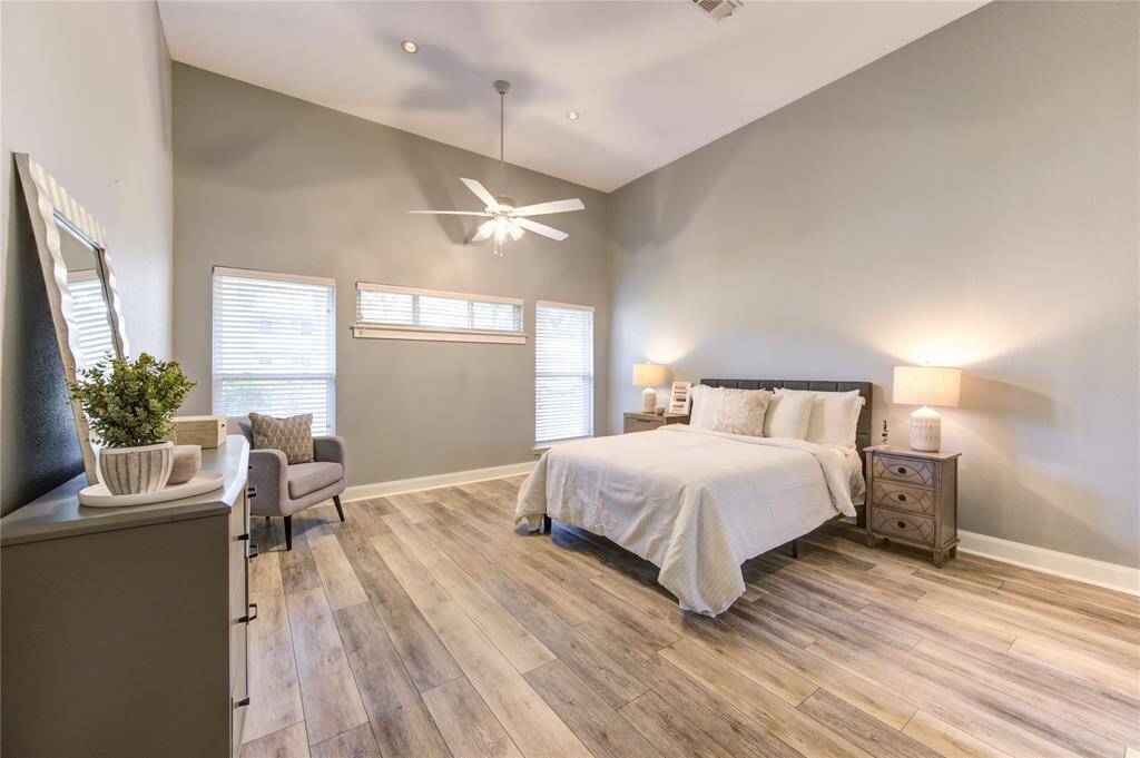 This spacious bedroom features a high ceiling with a ceiling fan, large windows for natural light, and stylish wood flooring. It includes a cozy sitting area and modern furnishings, creating a serene and inviting atmosphere.