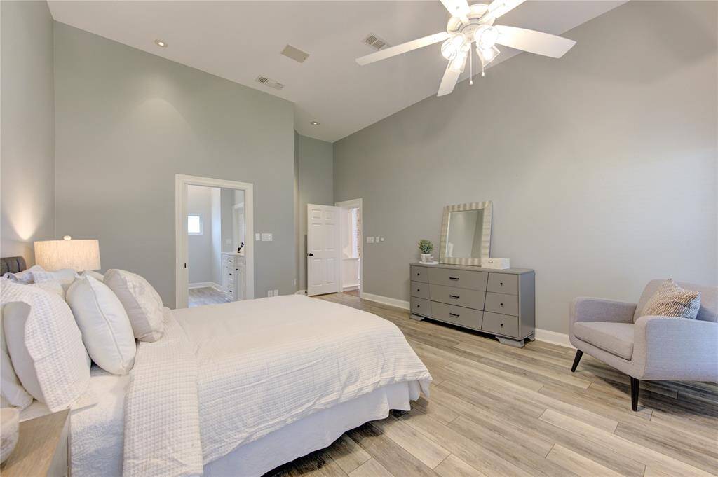 Spacious bedroom with high ceilings, modern light fixture, and neutral decor. Features wood-look flooring, a large dresser, and cozy seating. Includes an en-suite bathroom for convenience.