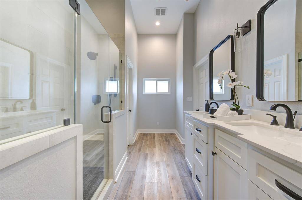 This modern bathroom features a sleek double vanity with black fixtures, a spacious glass-enclosed shower, and elegant wood flooring, creating a bright and sophisticated space.