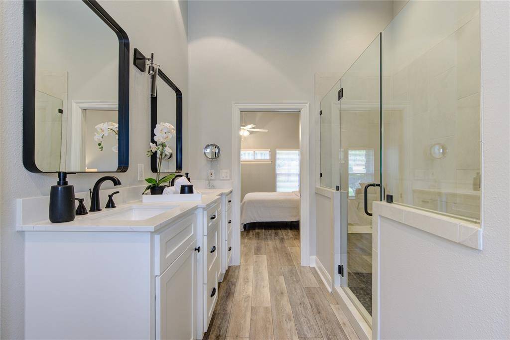 This photo showcases a modern bathroom featuring a double vanity with sleek black fixtures, a spacious glass-enclosed shower, and light wood flooring. The room flows seamlessly into a bright bedroom, creating an open and airy feel.
