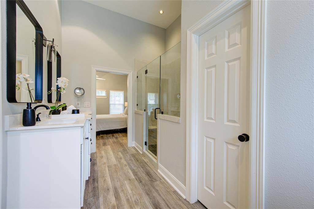 This photo shows a bright, modern bathroom featuring a spacious glass shower, dual vanity with stylish mirrors, and sleek wood-like flooring. The room leads into a cozy bedroom, creating a seamless transition between the spaces.