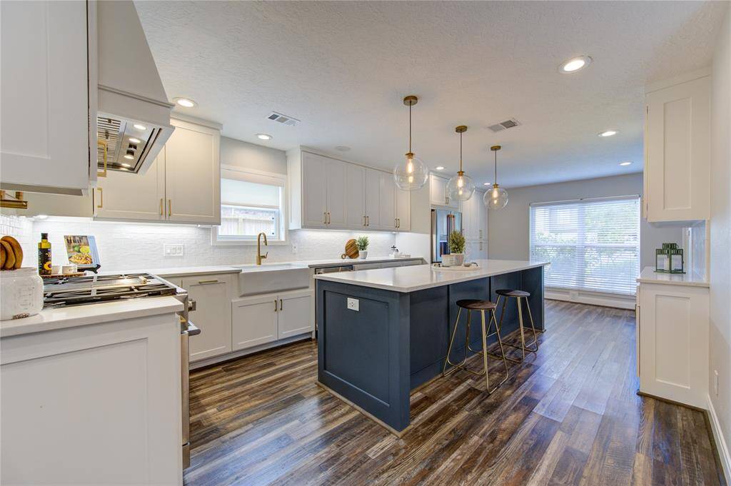This modern kitchen features sleek white cabinetry, a large navy island with seating, and stylish pendant lighting. It includes stainless steel appliances, a farmhouse sink, and wood-look flooring. Large windows provide ample natural light.