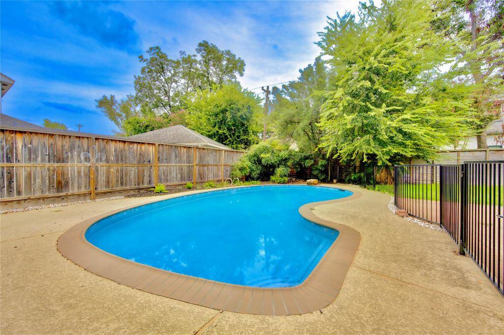 This backyard features a spacious, kidney-shaped pool surrounded by a concrete deck. It is enclosed by a wooden fence, with lush greenery providing a sense of privacy and tranquility.