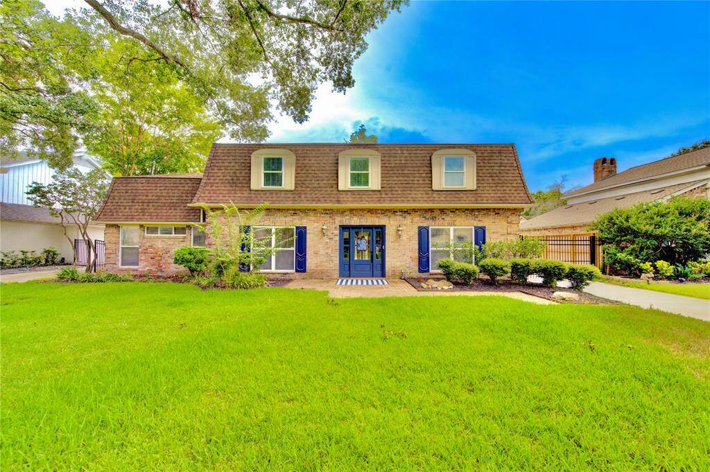 Charming two-story brick home with a distinctive gambrel roof, featuring large front windows and manicured landscaping. The spacious front lawn and inviting blue front doors enhance its curb appeal.