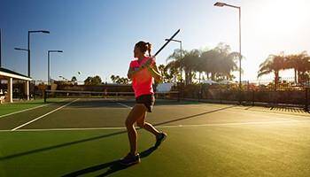 The photo shows a sunlit tennis court with a player in action, surrounded by palm trees, suggesting a vibrant community with recreational facilities.