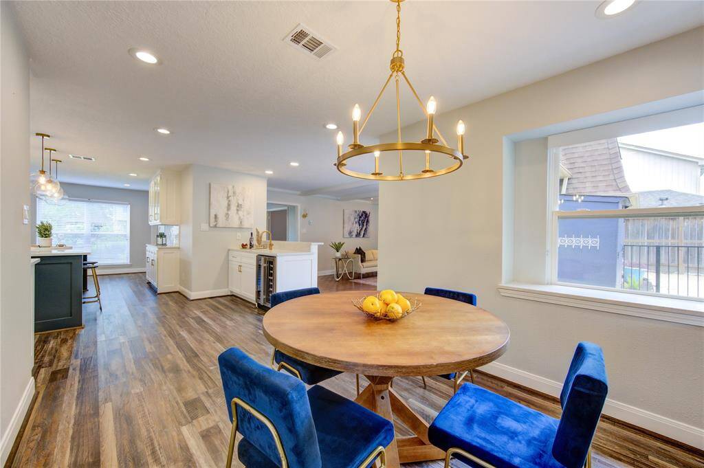 This spacious dining area features a round wooden table with elegant blue chairs, a stylish chandelier, and large windows for natural light. The open layout includes a view into the modern kitchen and living space, highlighted by wood flooring and recessed lighting.