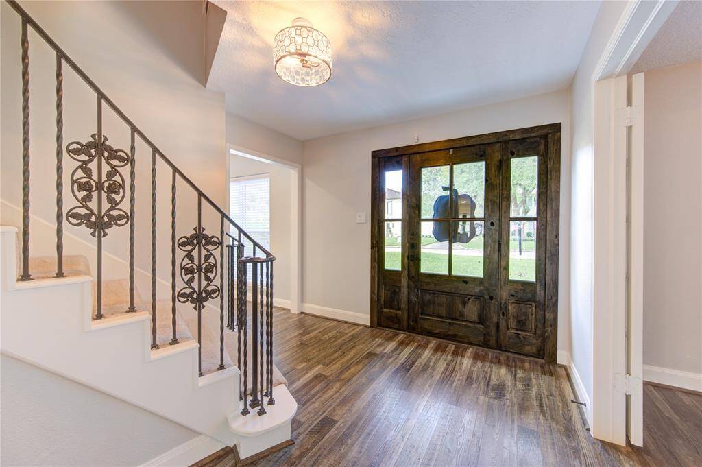 This photo showcases a bright entryway featuring a rustic wooden front door with large glass panels. The space has elegant wooden flooring and a staircase with intricate wrought iron railing details. A modern light fixture adds a touch of sophistication.