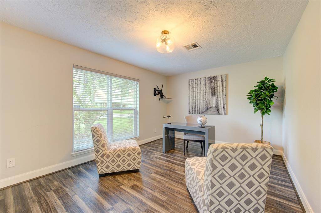 This room features large windows providing natural light, stylish wood flooring, and neutral walls. It includes a modern desk and seating area, ideal for an office or study space.