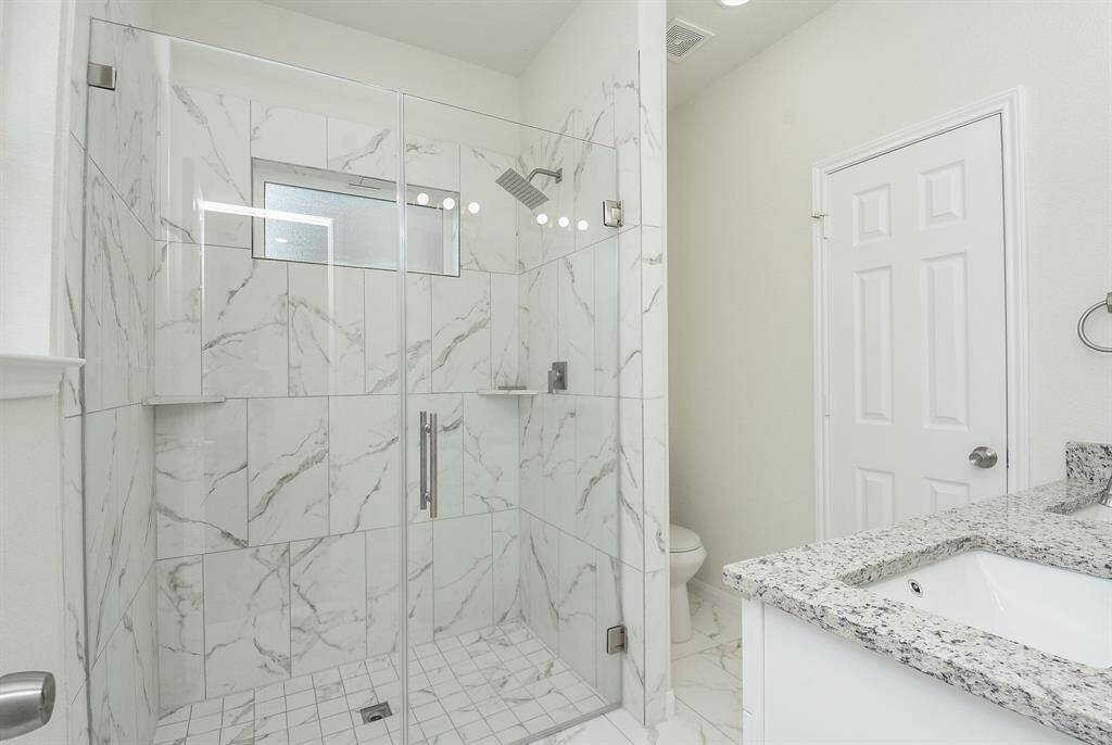 This is a modern bathroom featuring a large walk-in shower with marble-like tiles and glass doors, complemented by a sleek showerhead and built-in shelving. The space is bright and airy, with natural light coming from a window.