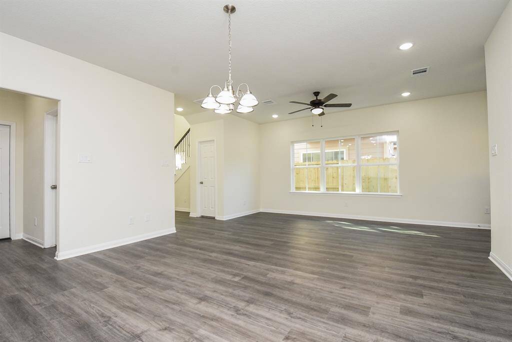 Spacious, well-lit room with modern dark wood flooring, neutral walls, elegant lighting fixtures, and large windows overlooking the backyard. The open layout provides a versatile space for living and dining areas.