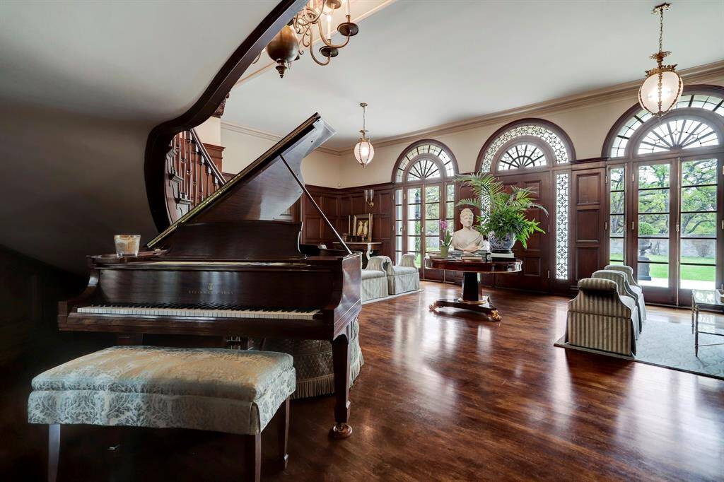 View of the opulent grand reception hall from under the stairs.  The rich wood paneled walls and hardwood floors gleam from the abundance of natural light that floods through the triple arch windows and front door with fan light.