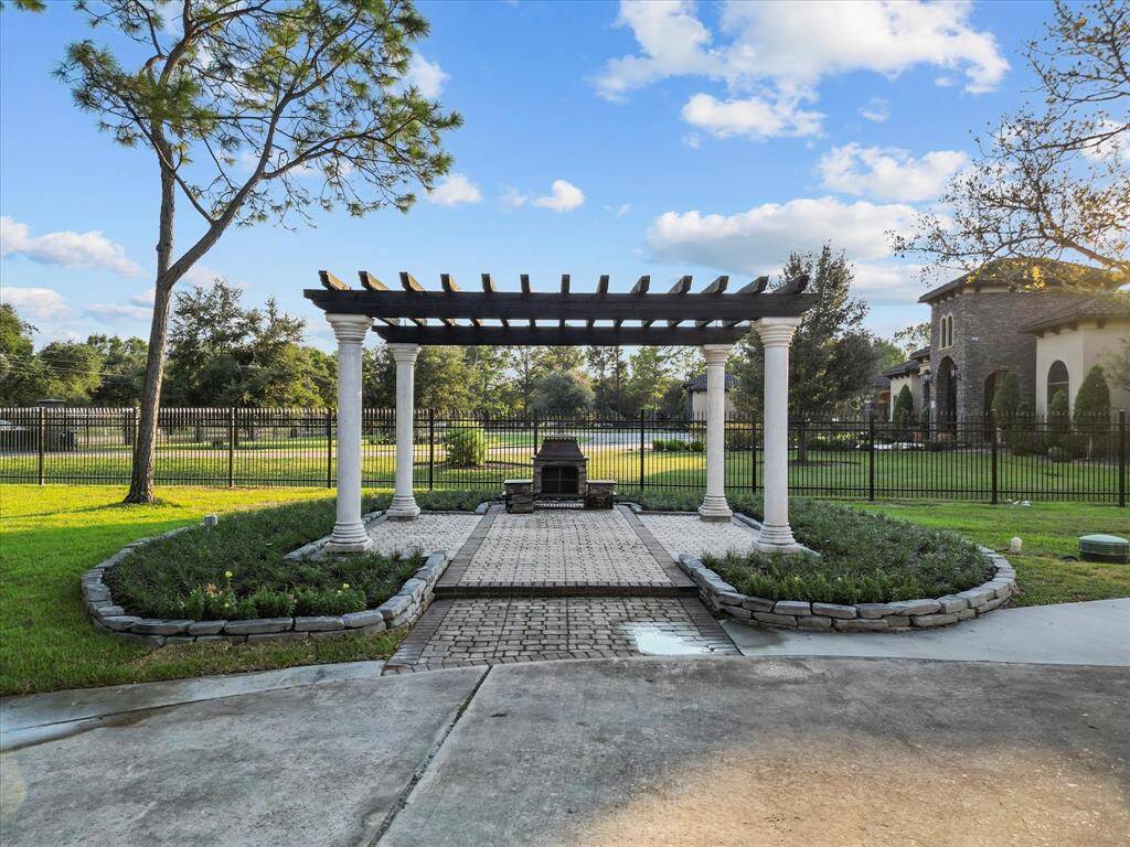 Elegant outdoor space featuring a pergola with classic columns, well-manicured landscaping, and a backdrop of a stately home and lush lawn, enclosed by a wrought-iron fence.