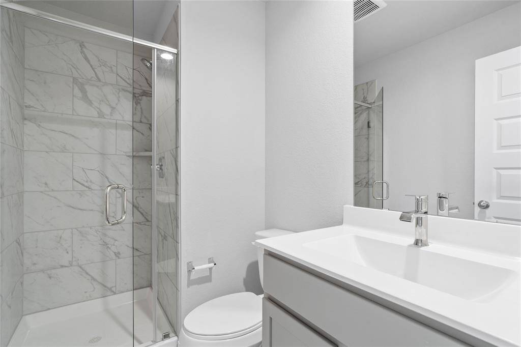 This private bath features tile flooring, shower with tile surround, stained wood cabinets, beautiful light countertops, mirror, dark, sleek fixtures and modern finishes.