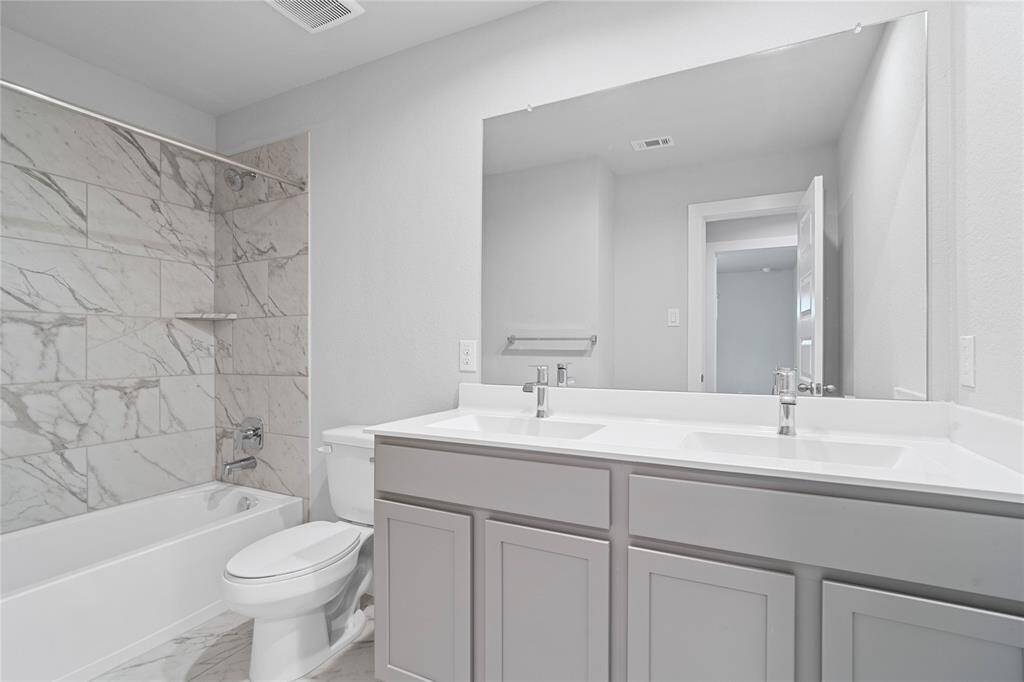Secondary bath features tile flooring, bath/shower combo with tile surround, stained wood cabinets, beautiful light countertops with dual vanities, mirror, dark, sleek fixtures and modern finishes.