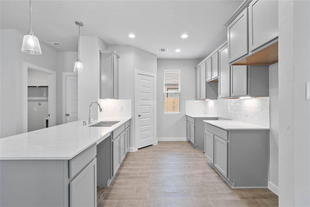 Another view of this beautiful kitchen showcasing the quartz countertops, stainless steel single bowl sink, under cabinet lighting, modern tile backsplash, pendant lighting, and a walk-in pantry.