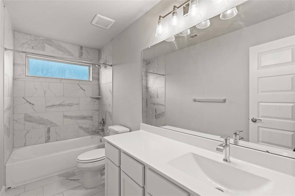 Secondary bath features tile flooring, bath/shower combo with tile surround, cool stained wood cabinets, beautiful light countertops, mirror, dark, sleek fixtures and modern finishes.