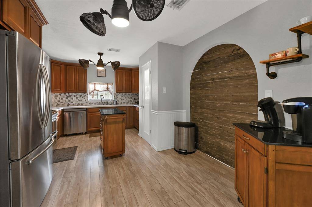 The beautifully updated kitchen features stainless steel appliances, wood cabinetry, a tiled backsplash, and wood like flooring. It also has a unique arched wooden accent wall, modern lighting fixtures, and a small island.