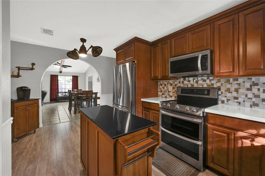 This is a spacious kitchen with rich wooden cabinetry, modern stainless-steel appliances, a central island, and a tile backsplash. The flooring is wood-look tile, and there's an arched opening leading to the dining area with ample natural light.