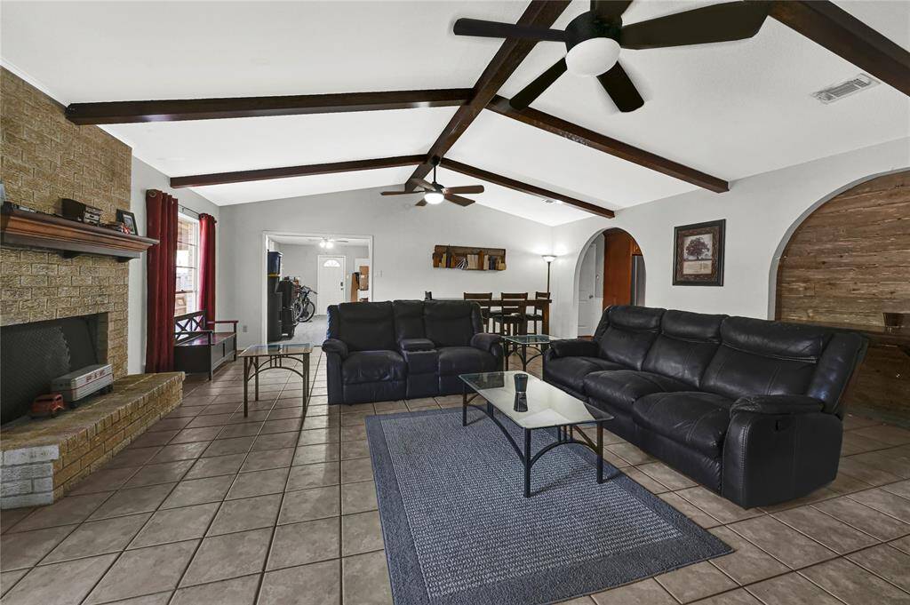 Spacious living room featuring a large stone fireplace, tile flooring, exposed wooden beams, and a vaulted ceiling with a ceiling fan.