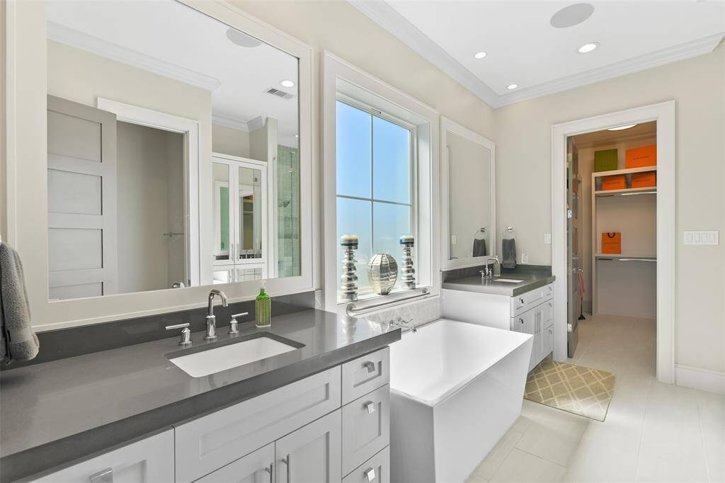 This expansive and luxurious primary ensuite bathroom features a freestanding tub, sleek quartz countertops with undermount trench-style sinks, and a thoughtfully designed vanity. The vanity is split by the tub, offering one side with generous counter space and the other with a more compact layout, both adorned with elegant framed mirrors.