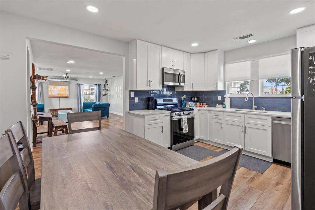 This is a bright, modern kitchen with white cabinetry, stainless steel appliances, dark blue subway tile backsplash, and a dining area with a wooden table and chairs. The room features ample natural light and wood flooring, creating a warm and inviting space.