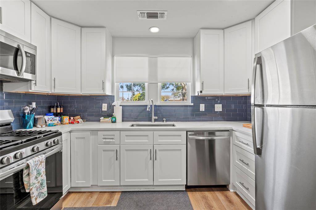 The photo showcases a modern, open-plan kitchen with white cabinetry and dark blue backsplash, stainless steel appliances, and ample natural light. An adjoining dining space seamlessly connects to a cozy living area, creating a spacious and inviting atmosphere for entertaining and daily living.