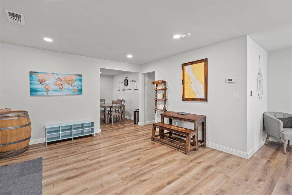 This is a bright and open space featuring modern wood-look flooring, neutral wall colors, recessed lighting, and tasteful decor. The area is versatile, currently showcasing a cozy nook with a bench, a decorative barrel, and a world map artwork, leading into a dining space with a view into another room.