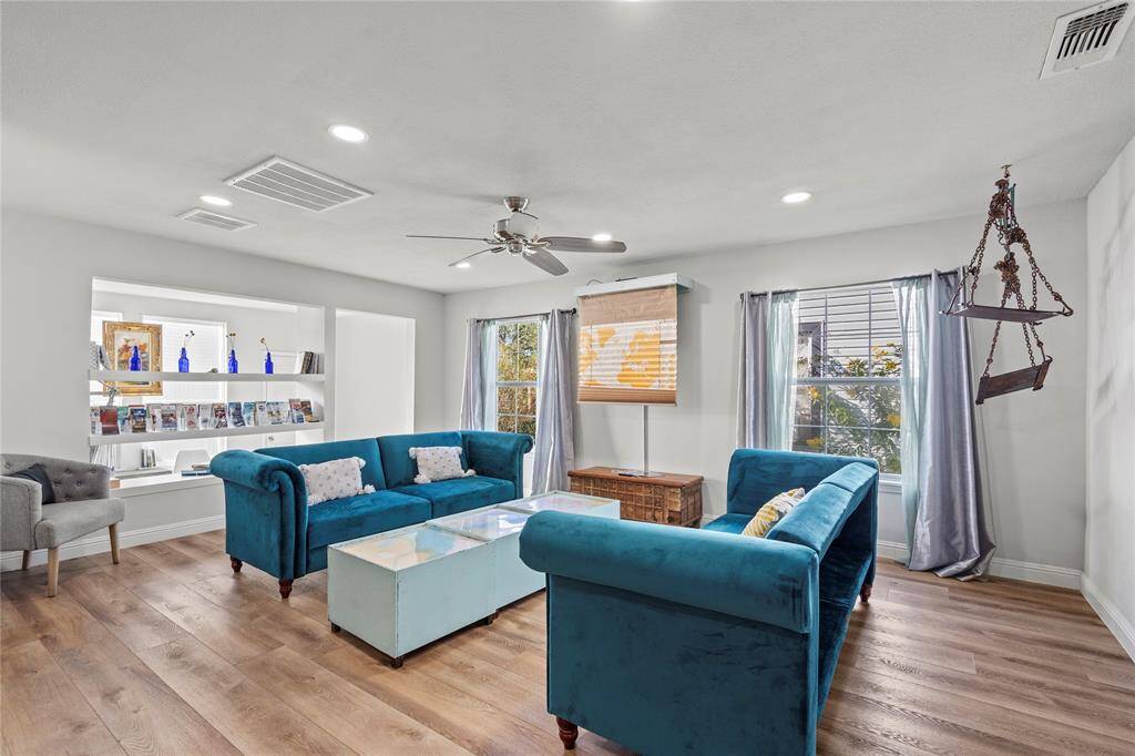 This is a bright, modern living room featuring elegant floors, recessed lighting, and a chic blue sofa set. There's a unique built-in shelving unit for displays, and large windows draped with sheer curtains allow for plenty of natural light. A distinctive hanging swing chair adds a touch of whimsy to the space.