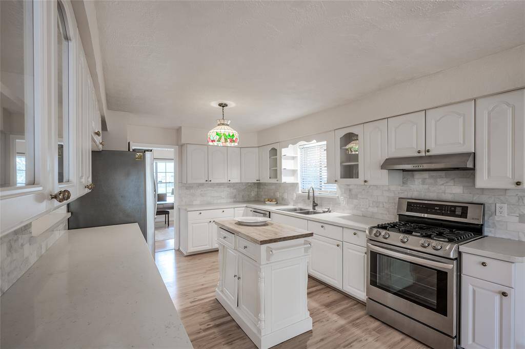 This is a bright, modern kitchen featuring white cabinetry, stainless steel appliances, butcher block island, a marble subway tile backsplash, and wood flooring. A decorative stained-glass pendant light adds a touch of character.