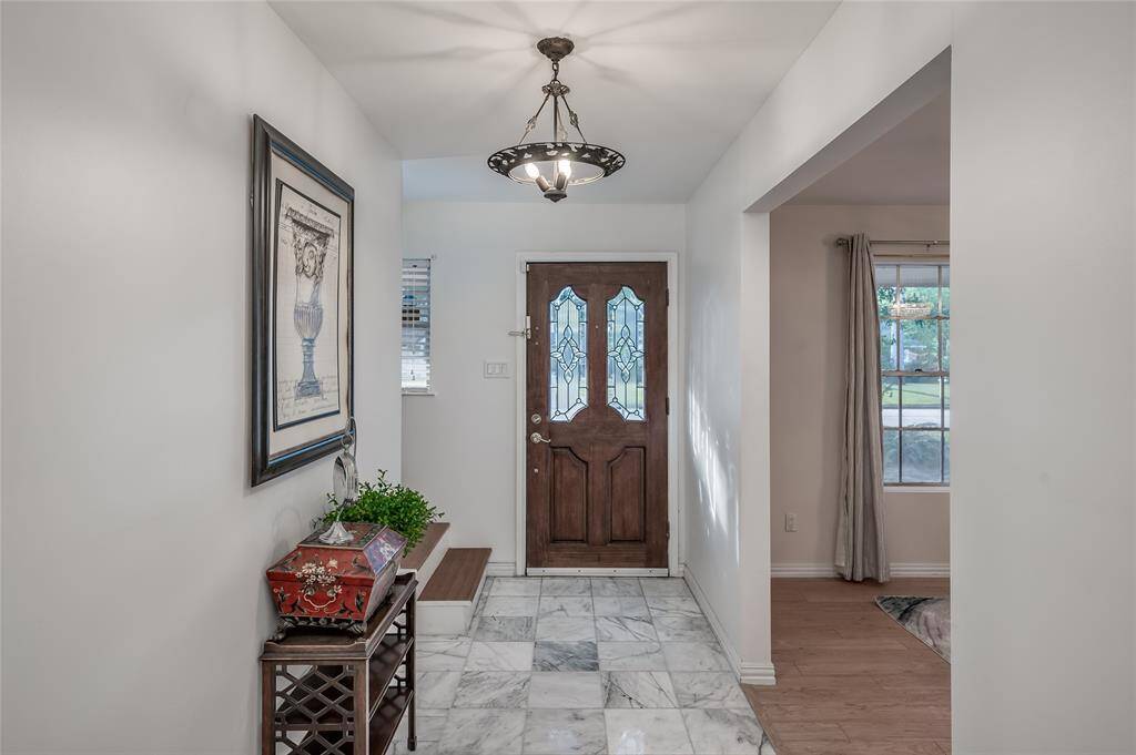 This is a well-lit entryway featuring a wooden front door with decorative glass, marble flooring that transitions into light engineered hardwood flooring, elegant light fixtures, and neutral wall colors, creating a welcoming atmosphere.