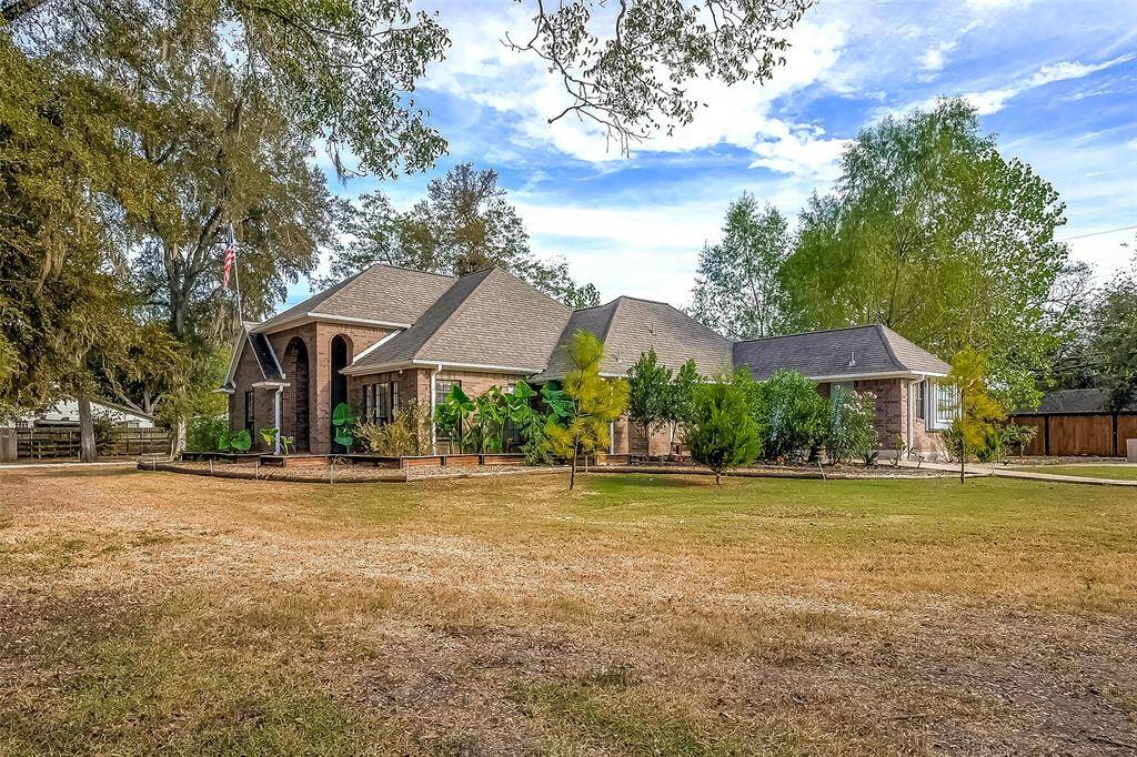 This is a spacious single-story home featuring a large front yard with mature trees, an expansive driveway, and a welcoming entryway. The property offers ample outdoor space and a detached structure that could serve multiple purposes.