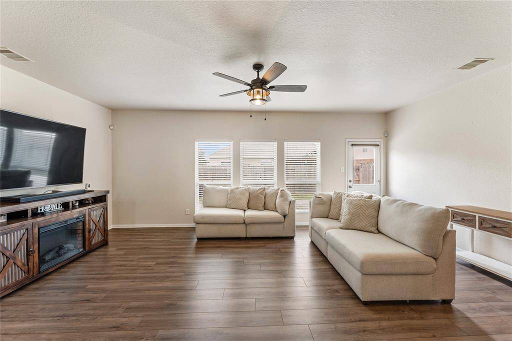 This Is a Spacious And Well-lit Living Room Featuring Contemporary Wood Flooring, Neutral Wall Colors, A Ceiling Fan, And Ample Natural Light From Windows With Blinds. Access To Outdoor Space Is Provided Through A Glass Door.