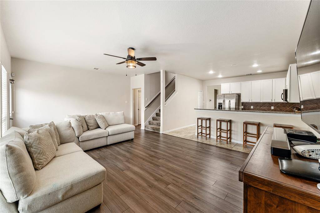 This Is A Modern, Open-concept Living Space Featuring A Comfortable Living Area With A Large Sectional Sofa And Hardwood Flooring, Leading Into A Kitchen With White Cabinets, Stainless Steel Appliances, And A Breakfast Bar. The Room Is Well-lit With Natural Light And Includes A Ceiling Fan.