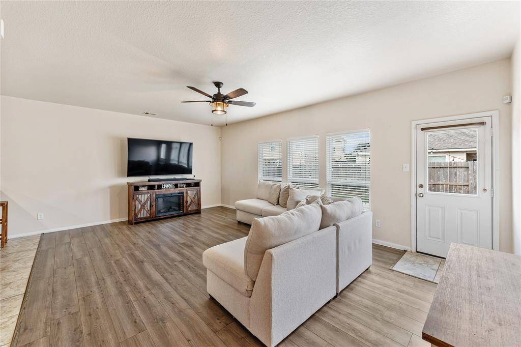 A Bright, Spacious Living Room With Modern Wood Flooring, Able To Fit A Large Comfortable Sofa And  Flat-screen TV, Ample Natural Light From Windows, And a Ceiling Fan.