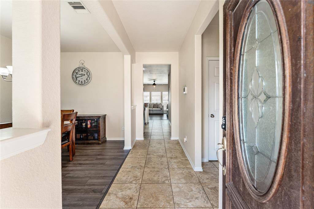 This Is A Well-lit Entryway With tiled Floors Leading Into A Spacious Interior. The Front Door Features Decorative Glass And A Wooden Finish, And There's a Glimpse Of A Cozy Dining Room. The Neutral Color Palette Provides A Warm And Inviting Atmosphere.