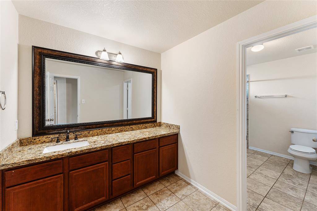 This is a neat and well-maintained bathroom featuring a large vanity with granite countertop and ample cabinet space, a wide mirror, and tiled flooring. There's a separate toilet area visible through the doorway.