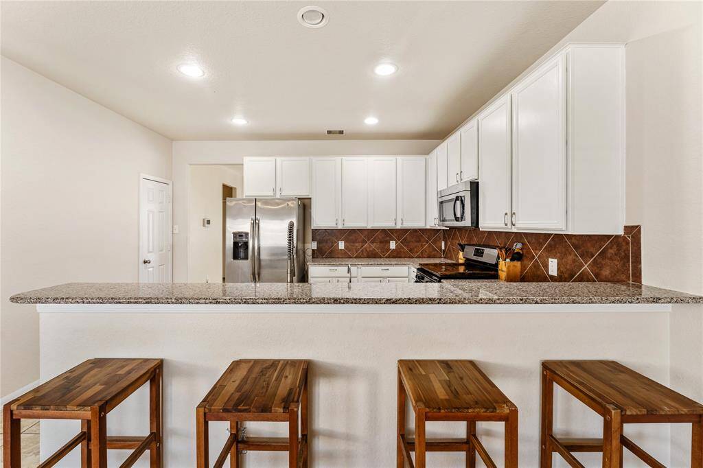 This Is A Modern Kitchen With White Cabinetry, Stainless Steel Appliances, Granite Countertops, And A Tile Backsplash. It Includes A Breakfast Bar With Seating.