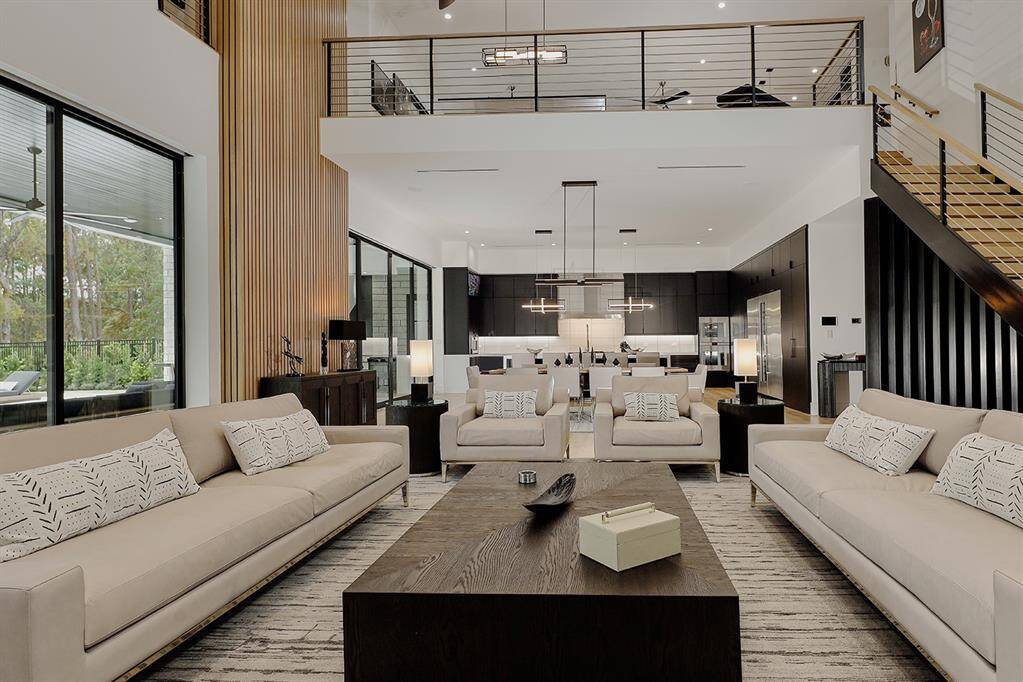 Another view of open-concept living space with high ceilings, large windows offering natural light, and a sleek kitchen visible in the background. The area features a neutral color palette and a second-floor balcony with a glass railing overlooking the living room.