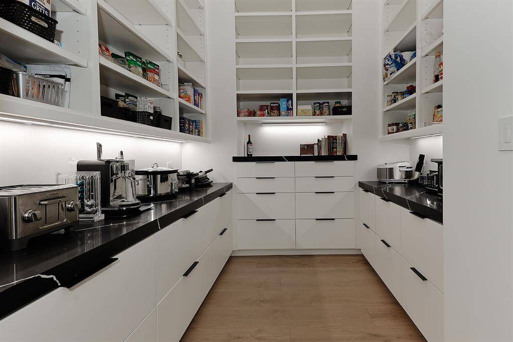 Spacious well-organized walk-in pantry with ample shelving and storage space, black quartz countertops.  Equipped with under-shelf lighting  all set on a warm wooden floor.