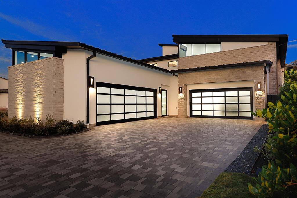 Amazing home featuring clean lines, a flat metal & TPO roof design, a spacious paver circular driveway leading to four car double garage with stylish frosted glass doors. The exterior is illuminated by sleek wall sconces, enhancing the home's upscale facade during the evening hours. Liftmaster side entry commercial garage doors. Workshop & AC storage.