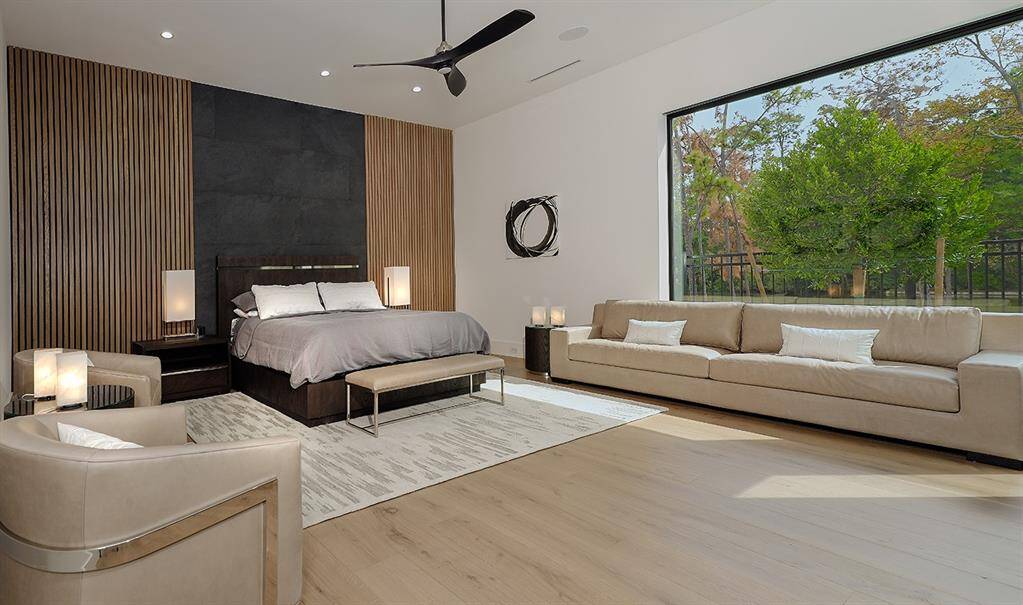This modern bedroom features a large bed with a stylish headboard against a textured wall, flanked by wooden paneling. It has an expansive floor-to-ceiling window offering garden views, a cozy seating area, and a soft, neutral color palette enhancing the room's serene ambiance.