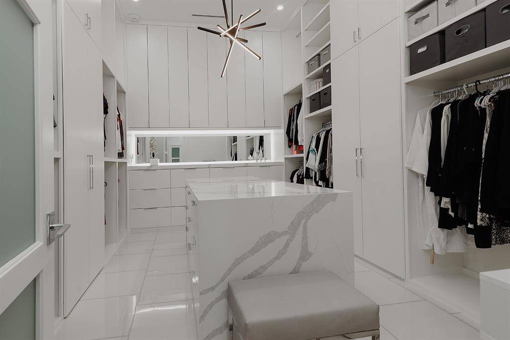 This is a spacious, modern walk-in closet featuring white custom cabinetry, a center island with quartz waterfall countertop and stylish light fixture overhead. There's ample storage with both open shelving and soft closing drawers, providing a clean and organized space for clothing and accessories.