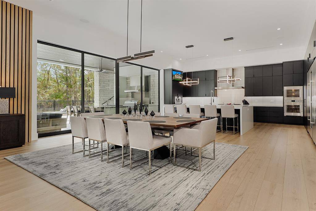 This is a modern open-plan living space featuring a sleek kitchen with black cabinetry and integrated appliances, a dining area with a large table and comfortable chairs, and an inviting living area with ample natural light from large windows leading to an outdoor space. The design is complemented by light wood flooring and contemporary lighting fixtures.