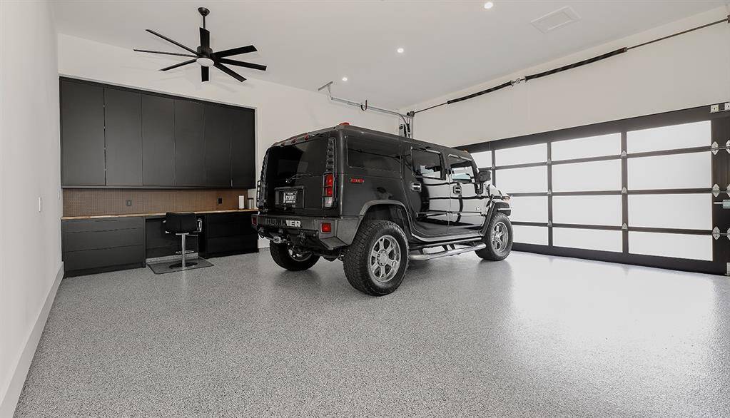 This is a spacious, modern garage featuring a clean, epoxy-coated floor, large storage cabinets, a workbench area, and a contemporary ceiling fan/light fixture. The garage door is frosted glass with black frames, allowing for natural light while maintaining privacy.   Liftmaster side entry commercial garage doors. Workshop & bench. Two EV outlets. Easy attic pull down stairs for additional storage.