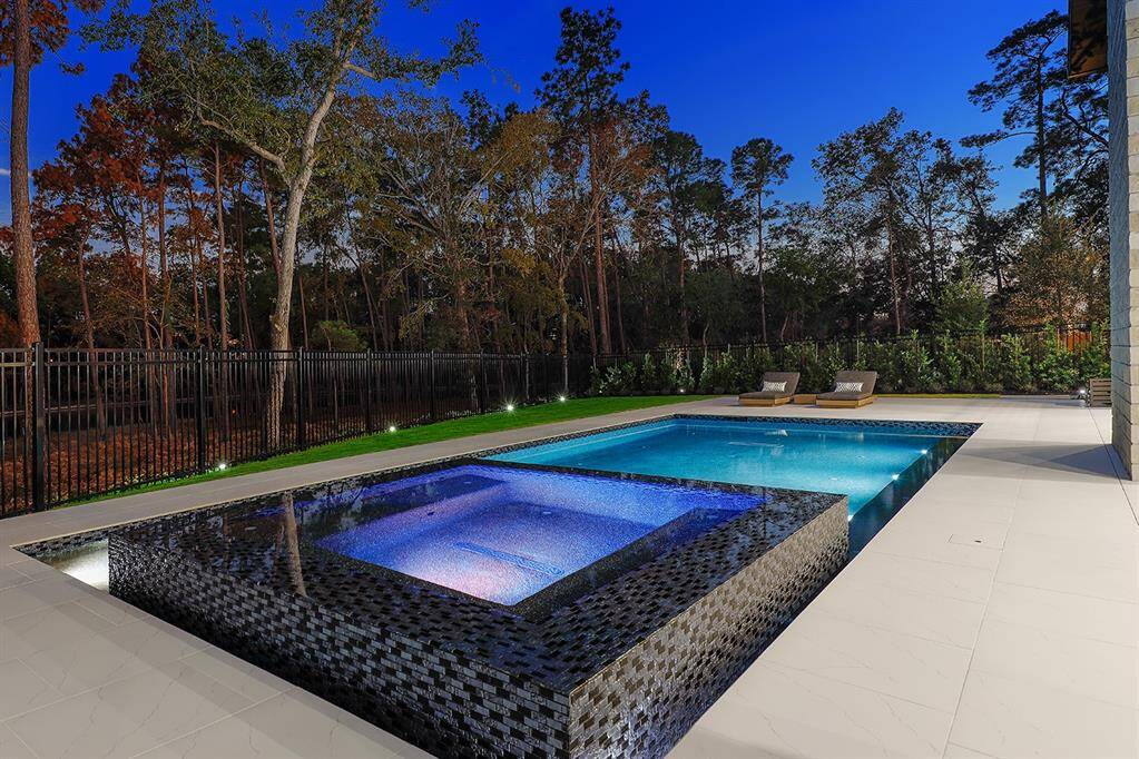 This is an elegant backyard featuring a modern rectangular pool with an attached spa, set against a backdrop of tall, mature trees and view of Lake Woodlands. The area includes a spacious tiled patio, perfect for relaxation. The property is enclosed with a black wrought iron fence, ensuring privacy and safety.