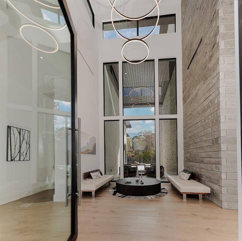 This is a spacious foyer featuring 26' high ceilings, large windows for ample natural light, sleek furnishings, and a blend of natural textures with stone walls and wood flooring. The room opens to an outdoor view, offering a seamless indoor-outdoor living experience.